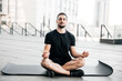 © Ivan - Man practicing yoga in big city. Sporty man meditating after workout. Excercising at home. Young man with stubble sitting on black yoga mat with eyes closed outdoors and relaxing in easy seat pose.