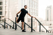 © Ivan - Athletic man stretching legs before workout outdoors. Runner in black sportswear excercising at morning holding handrail. Active living. Morning city on background. Morning warm up.