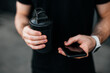 © Ivan - Close up cut view male hands using smartphone and holding sports bottle. Resting time Runner in black t-shirt. Modern technologies concept. Urban sport. Gray background.
