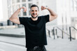 © Ivan - Cheerful man showing his muscles. Hard workout concept. Fit man showing biceps and smiling. Gray morning city on background. Handsome man excercised a lot to get a fit body.
