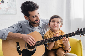 Hispanic father smiling while looking at son touching acoustic guitar, two generations of men