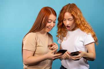 Surprised girlfriends on a blue background looking excited in the smartphones. People lifestyle concept.