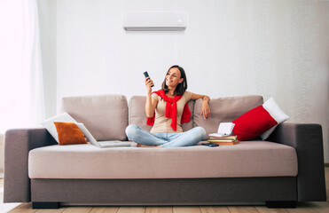 Beautiful modern brunette woman is using the air conditioner while sitting on the couch and resting in the hot day