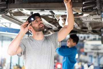 Wall Mural - automotive mechanic man using flash light checking under car damage at auto repair garage on lifter hoist with assistant. diversity people work together. vehicle maintenance. after service concept