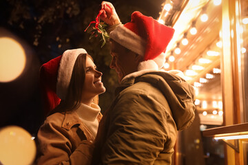 Poster - Happy couple in Santa hats standing under mistletoe bunch outdoors, bokeh effect