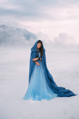 Art photo. Fantasy young woman fairy elf in blue cape with hood stands in cold wind. Winter nature background, mountains in the clouds, dramatic sky white snow. Girl Queen walks in dress, silk cloak