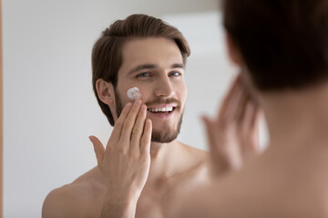 Smiling handsome millennial caucasian man looking in mirror, moisturizing face with nourishing creme, feeling satisfied with smoothed skin condition, enjoying morning cleansing routine in bathroom.