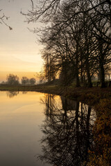  Orange glow in a swamp land landscape with trees and remains of Nijenbeek castle reflecting and mirrored in the still water in The Netherlands along the river IJssel at sunrise