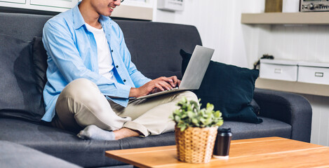 Wall Mural - Handsome asian businessman looking at technology of laptop computer monitor while sitting on sofa.Young creative coworkers business people working and typing on keyboard at home