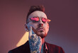 © Fxquadro - Serious and tattooed hipster businessperson with beard and stylish haircut poses in dark room with lights and colourful walls touching his face with finger.