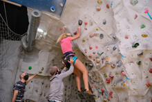 Group Of Three Young Friends Wearing Protective Face Masks Climbing. Woman Falling And Man Touching Her Butt On Artificial Rock Climbing Wall Indoors. Extreme Sports In The New Normal.