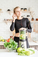 Wall Mural - Young blond smiling woman in black sportwear listening to the music and making green smoothie at home kitchen