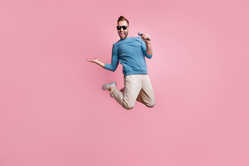 Wall Mural - Full length photo of handsome cute guy wear blue pullover black glasses jumping singing holding arm isolated pink color background