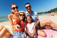 Family, Leisure And People Concept - Happy Mother, Father And Two Daughters Having Picnic On Summer Beach And Taking Picture With Selfie Stick