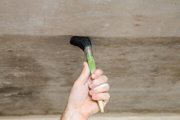 Young adult man hand holding brush and applying primer on concrete ceiling. Closeup. Repair work of home.