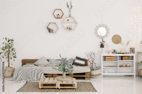 White living room. A sofa made of wooden pallets, green plants in pots, a chest of drawers, a round mirror in a boho style, shelves against a white wall. Scandinavian interior style. Minimalism