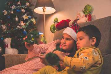 Escena infantil navideña. Niño y niña sentados en el sofá con el árbol de Navidad detrás juegan con sus juguetes. Infancia feliz de dos hermanos.