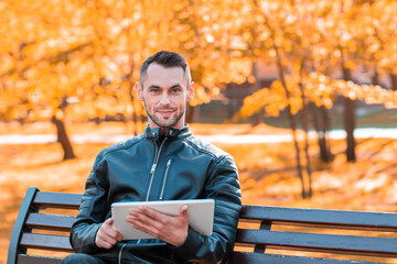 Handsome Young Man Sitting on the Bench and Using Big White Tablet PC at the Beautiful Autumn Park