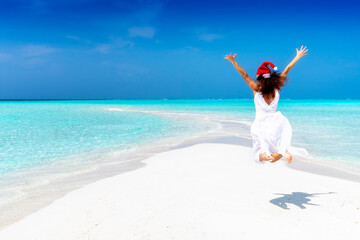 Christmas vacation concept with a woman in white dress and santa claus hat jumping on a tropical paradise beach with turquoise sea and fine sand