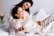 Hispanic lesbian woman hugging cheerful partner in pajamas on bed at morning