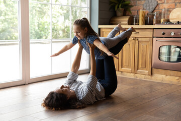 Cute gymnastics. Happy smiling young mom supporting little daughter in air doing flying plane exercise. Grown elder sister playing active game with younger girl lying on heated floor at home kitchen