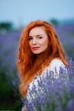Fototapeta Lawenda - Summer portrait of a beautiful girl with long curly red hair. European girl in lavender field. Wavy Red Hair