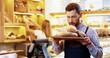 © VAKSMANV - Close up of happy bearded young Caucasian man seller in apron standing in bakery and holding tray with fresh baked bread in hands, smelling baking and looking at camera while colleague works behind