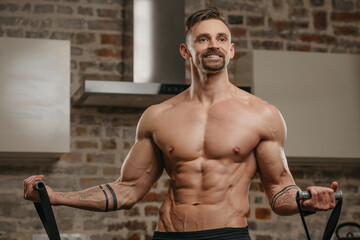 Wall Mural - A muscular man is working out with an elastic pull rope in his apartment. Athletic guy with tattoos on his forearms is demonstrating his sporty physique at home. A smiling athlete with a naked torso