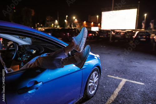 Close up of woman s legs dangling out a car window parked in front of a big white screen at drive in cinema