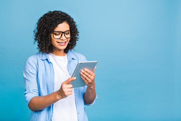 Portrait of young american student girl with curly african hair holding digital tablet and smiling standing over isolated blue background with copy space for text, logo or advertising.