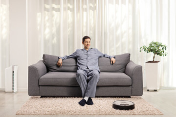 Canvas Print - Man in pajamas resting on a sofa at home with a robotic vacuum cleaner