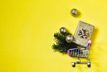 Christmas Creative Composition. Shopping Cart And Christmas Gold Gift, Gold Decorations, Fir Tree Branches On Yellow Background. Christmas, New Year, Winter Concept. Flat Lay, Top View, Copy Space.