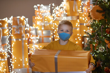 A little boy in medical masks holding a gift in his hands. He is near Christmas tree at home. Happy New Year Covid 2021