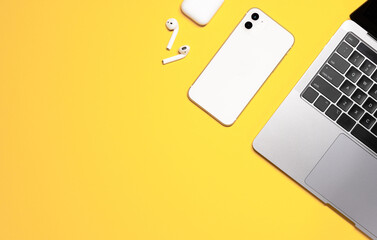 Colorful flat lay shot of a silver grey laptop computer, white smartphone, earphones with a charging case on a yellow background.