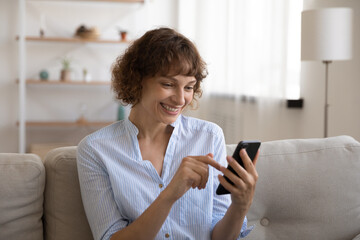 Happy young female rest on sofa at living room making video call by cell shooting selfie using phone webcam. Smiling woman enjoy chatting in app scrolling web pages reading electronic book good news