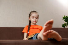 A Little Caucasian Girl With Bare Feet Reading Book While Sitting On A Sofa At Home. Selective Focus