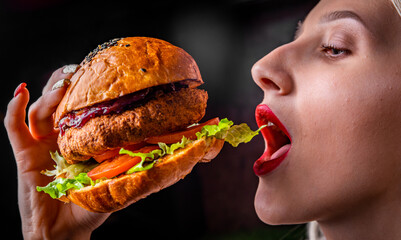 young woman eating a hamburger with hands over blurred black background