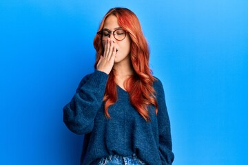 Poster - Young beautiful redhead woman wearing casual sweater and glasses over blue background bored yawning tired covering mouth with hand. restless and sleepiness.