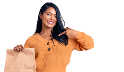 Hispanic woman with long hair holding take away paper bag pointing finger to one self smiling happy and proud