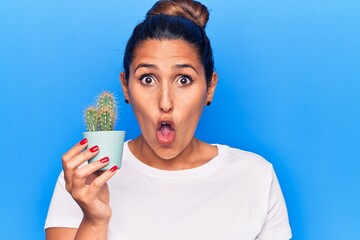 Sticker - Young beautiful brunette woman holding small cactus pot scared and amazed with open mouth for surprise, disbelief face