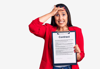 Poster - Young beautiful brunette woman holding clipboard with contract stressed and frustrated with hand on head, surprised and angry face