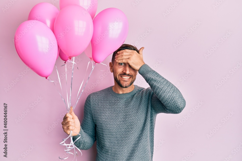 Handsome man with beard holding pink balloons stressed and frustrated with hand on head, surprised and angry face