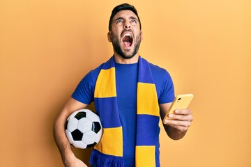 Young hispanic man football hooligan holding soccer ball using smartphone angry and mad screaming frustrated and furious, shouting with anger looking up.