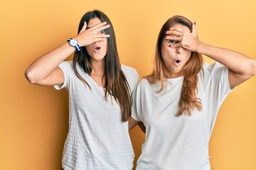 Poster - Hispanic family of mother and daughter wearing casual white tshirt peeking in shock covering face and eyes with hand, looking through fingers with embarrassed expression.