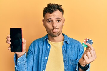 Young hispanic man holding poker chips and smartphone showing blank screen depressed and worry for distress, crying angry and afraid. sad expression.