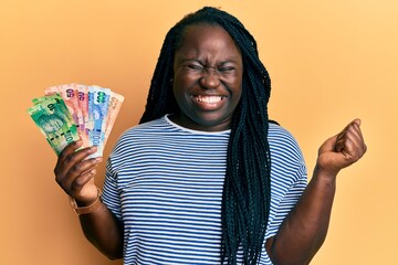 Young black woman with braids holding south african rand banknotes screaming proud, celebrating victory and success very excited with raised arm