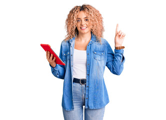 Young blonde woman with curly hair using touchpad device surprised with an idea or question pointing finger with happy face, number one