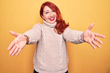 Poster - Young beautiful redhead woman wearing casual turtleneck sweater over yellow background looking at the camera smiling with open arms for hug. Cheerful expression embracing happiness.