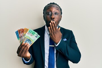 Poster - Handsome young black man wearing business suit and tie holding australian dollars covering mouth with hand, shocked and afraid for mistake. surprised expression