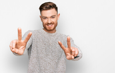 Young redhead man wearing casual winter sweater smiling with tongue out showing fingers of both hands doing victory sign. number two.